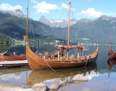 Several wooden Viking ships with dragon-headed prows are moored on a calm lake, with mountains and lush greenery in the background under a blue sky.