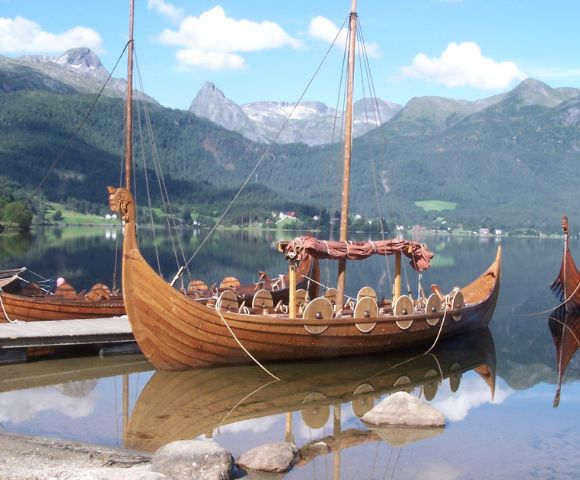 Several wooden Viking ships with dragon-headed prows are moored on a calm lake, with mountains and lush greenery in the background under a blue sky.