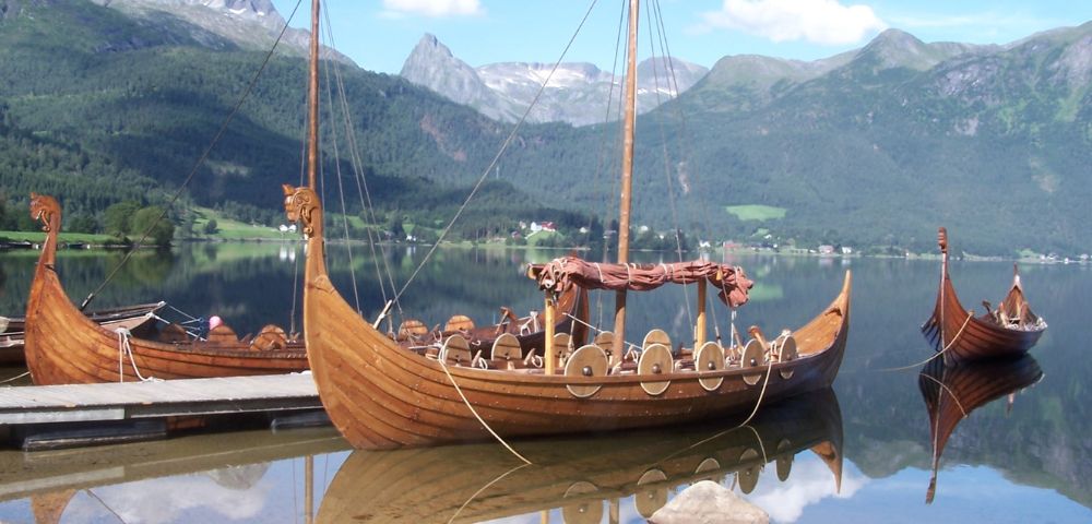 Several wooden Viking ships with dragon-headed prows are moored on a calm lake, with mountains and lush greenery in the background under a blue sky.