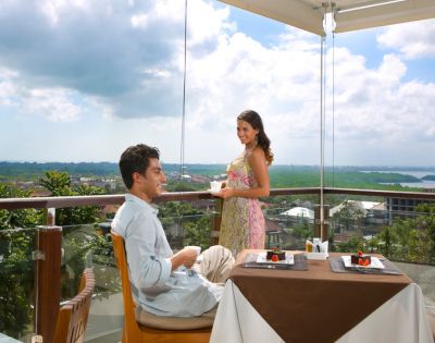A couple enjoys a meal on a sunny balcony overlooking a lush, green landscape. The man sits at a table with dessert, while the woman stands, smiling warmly.