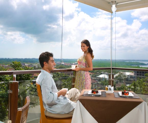 A couple enjoys a meal on a sunny balcony overlooking a lush, green landscape. The man sits at a table with dessert, while the woman stands, smiling warmly.