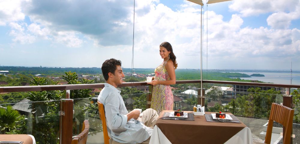 A couple enjoys a meal on a sunny balcony overlooking a lush, green landscape. The man sits at a table with dessert, while the woman stands, smiling warmly.