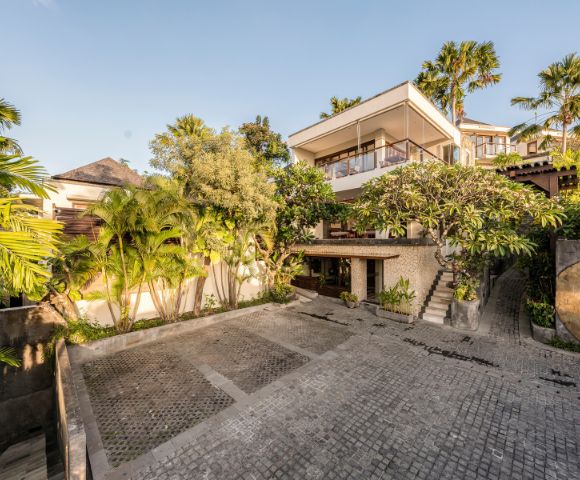 Luxurious hillside villa with a modern design, surrounded by lush palm trees and greenery. A serene courtyard with cobblestone paving adds an inviting touch.