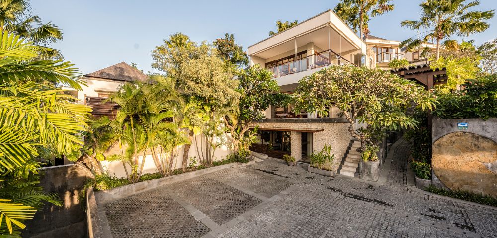 Luxurious hillside villa with a modern design, surrounded by lush palm trees and greenery. A serene courtyard with cobblestone paving adds an inviting touch.