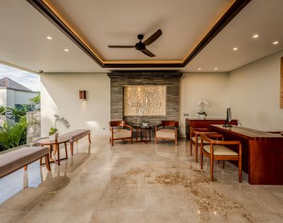 Spacious, elegantly decorated lobby with marble floor, wooden furniture, wall art, and ceiling fan. Sunlight filters through a large window, creating a serene ambiance.