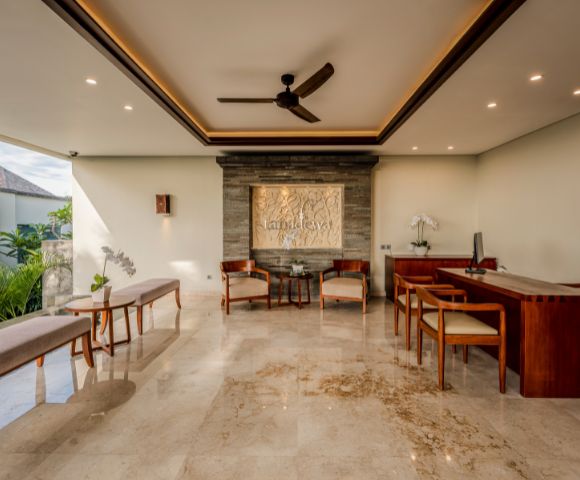 Spacious, elegantly decorated lobby with marble floor, wooden furniture, wall art, and ceiling fan. Sunlight filters through a large window, creating a serene ambiance.