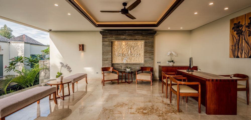 Spacious, elegantly decorated lobby with marble floor, wooden furniture, wall art, and ceiling fan. Sunlight filters through a large window, creating a serene ambiance.