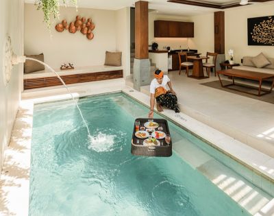 A person in a sunlit luxury villa places a floating breakfast tray on a private pool. A serene ambiance is enhanced by elegant interior decor.