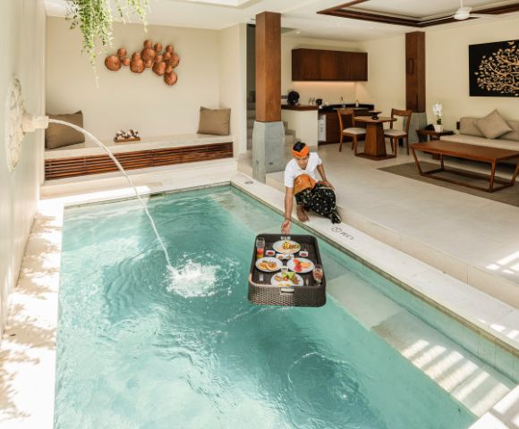 A person in a sunlit luxury villa places a floating breakfast tray on a private pool. A serene ambiance is enhanced by elegant interior decor.