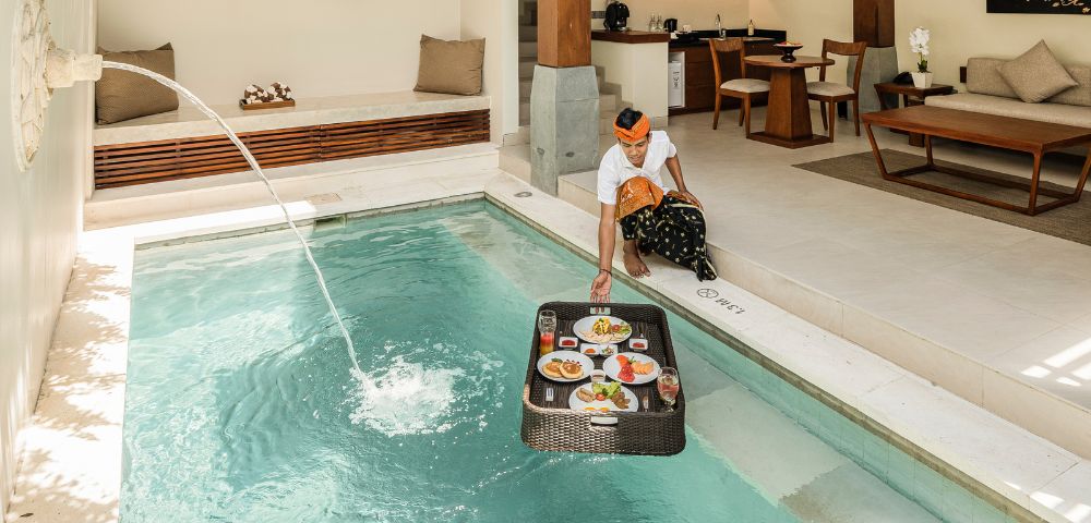A person in a sunlit luxury villa places a floating breakfast tray on a private pool. A serene ambiance is enhanced by elegant interior decor.