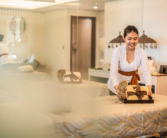 A spa attendant in a white uniform smiles while arranging towels on a massage table in a serene, warmly lit spa room, conveying a relaxing atmosphere.