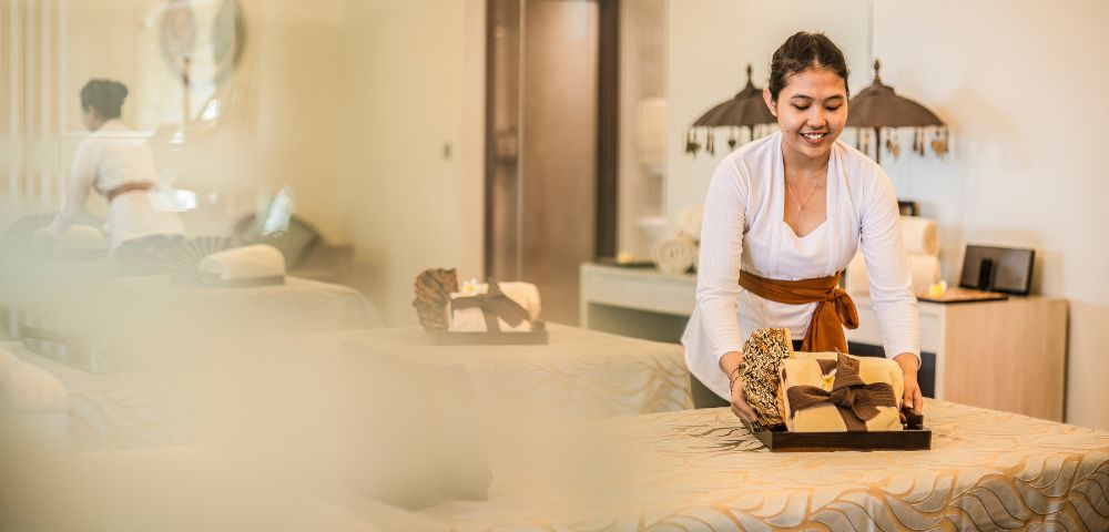 A spa attendant in a white uniform smiles while arranging towels on a massage table in a serene, warmly lit spa room, conveying a relaxing atmosphere.