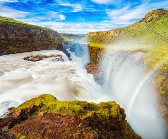 A majestic waterfall cascades through a lush green canyon, with mist creating a vibrant rainbow under a clear blue sky, conveying serenity and awe.