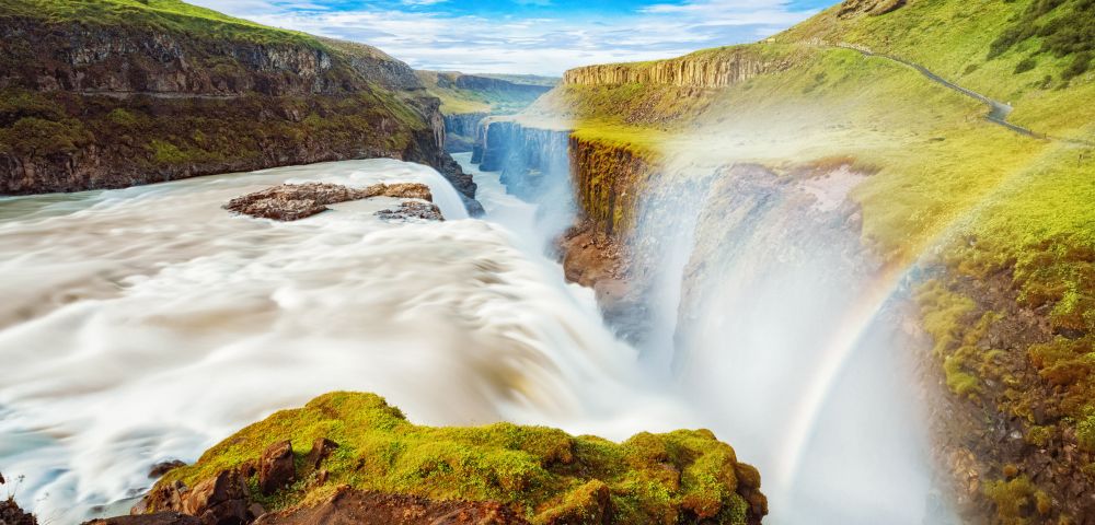 A majestic waterfall cascades through a lush green canyon, with mist creating a vibrant rainbow under a clear blue sky, conveying serenity and awe.
