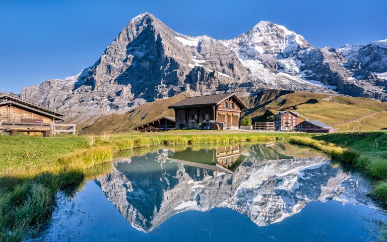 Wooden cabins sit by a tranquil pond, reflecting snow-capped mountains under a clear blue sky. The scene evokes serenity and natural beauty.