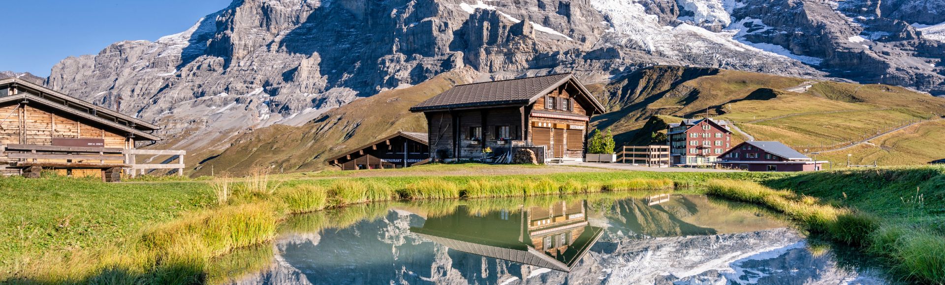 Wooden cabins sit by a tranquil pond, reflecting snow-capped mountains under a clear blue sky. The scene evokes serenity and natural beauty.