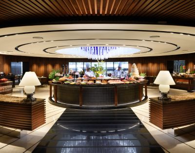 Elegant buffet restaurant with a modern, circular design. Central table filled with food, surrounded by warm lighting, decorative lamps, and wood accents. Cozy ambiance.