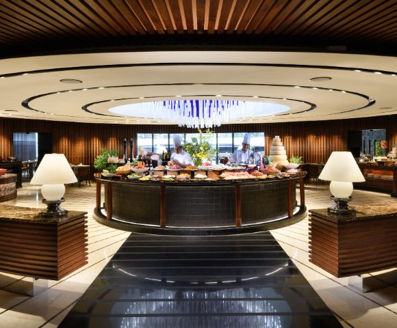 Elegant buffet restaurant with a modern, circular design. Central table filled with food, surrounded by warm lighting, decorative lamps, and wood accents. Cozy ambiance.