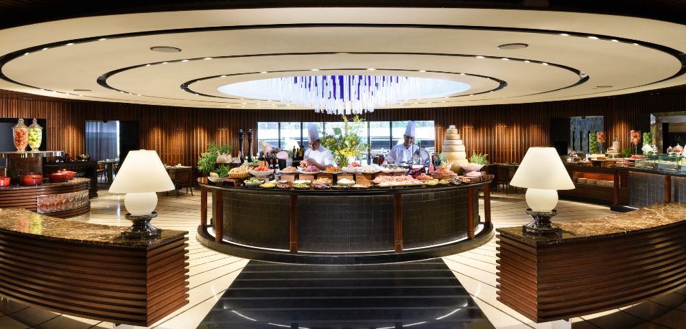 Elegant buffet restaurant with a modern, circular design. Central table filled with food, surrounded by warm lighting, decorative lamps, and wood accents. Cozy ambiance.