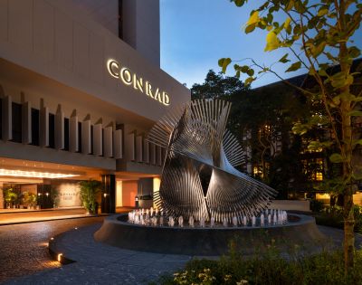 Modern hotel entrance at dusk featuring a large, intricate metal sculpture on a round fountain. The illuminated sign reads 