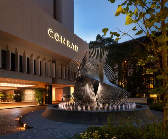 Modern hotel entrance at dusk featuring a large, intricate metal sculpture on a round fountain. The illuminated sign reads 