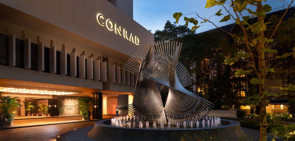 Modern hotel entrance at dusk featuring a large, intricate metal sculpture on a round fountain. The illuminated sign reads 
