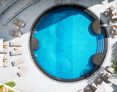 Aerial view of a circular swimming pool with clear blue water, surrounded by beige lounge chairs on a sunlit patio, creating a relaxing atmosphere.