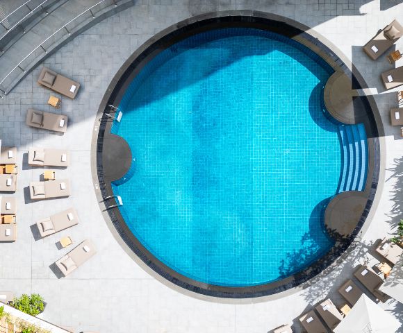 Aerial view of a circular swimming pool with clear blue water, surrounded by beige lounge chairs on a sunlit patio, creating a relaxing atmosphere.