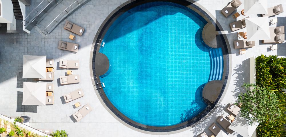 Aerial view of a circular swimming pool with clear blue water, surrounded by beige lounge chairs on a sunlit patio, creating a relaxing atmosphere.