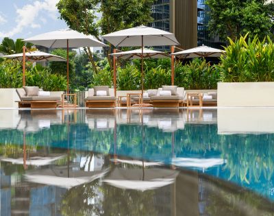 Poolside scene with plush loungers and umbrellas reflected in calm water. Surrounded by lush greenery, the setting evokes relaxation and tranquility.