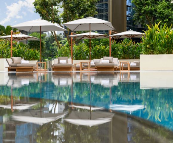 Poolside scene with plush loungers and umbrellas reflected in calm water. Surrounded by lush greenery, the setting evokes relaxation and tranquility.