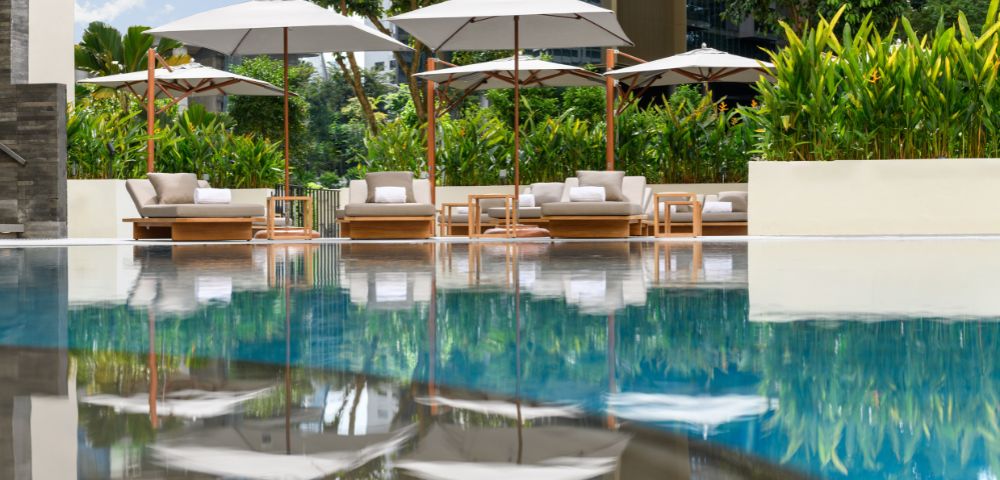 Poolside scene with plush loungers and umbrellas reflected in calm water. Surrounded by lush greenery, the setting evokes relaxation and tranquility.