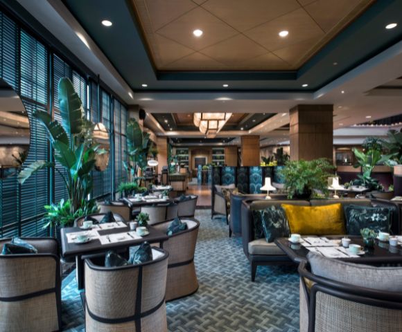 The elegant restaurant interior features wicker chairs, patterned carpets, and lush green plants, creating a cozy and inviting atmosphere.