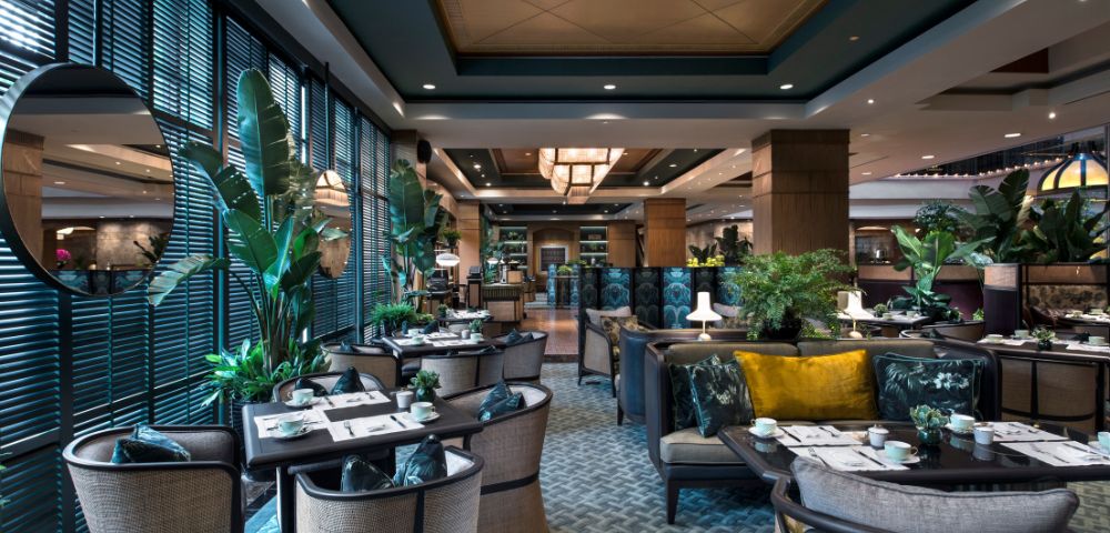 The elegant restaurant interior features wicker chairs, patterned carpets, and lush green plants, creating a cozy and inviting atmosphere.