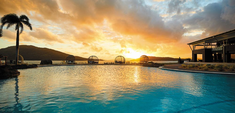 Sunset view over a tranquil pool with a silhouetted palm tree, lounge areas, and mountains in the background, creating a serene and warm atmosphere.