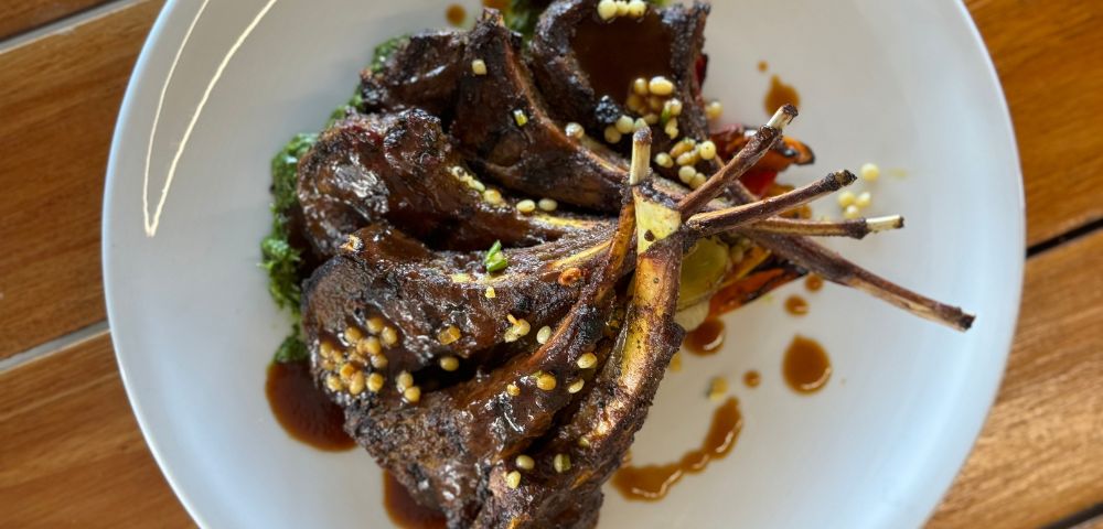 Grilled lamb chops with sauce and herbs on a white plate, garnished with grains, atop a wooden table. The dish looks savory and inviting.