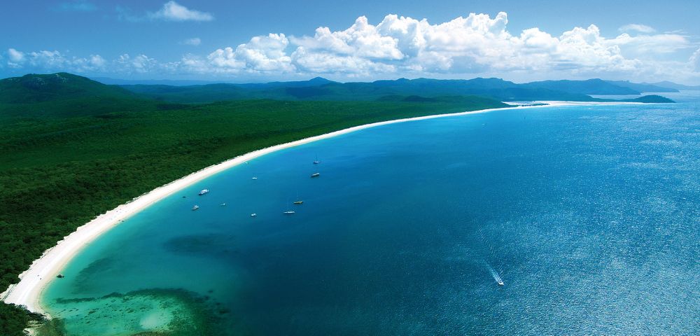 Expansive aerial view of a serene blue ocean meeting a curving white sandy beach, backed by lush green forest. Boats dot the clear, tranquil waters.
