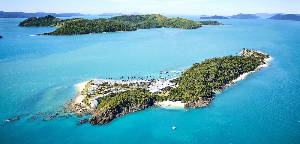 Aerial view of a serene tropical island with lush greenery, sandy beaches, and turquoise waters. Surrounding islands and clear skies enhance the tranquil scene.