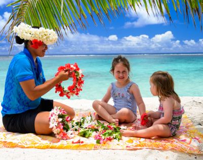 A woman in a blue shirt and floral headpiece making leis on a tropical beach with two young girls. They sit on a blanket under palm fronds, smiling.