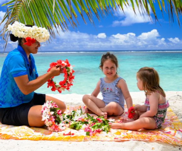 A woman in a blue shirt and floral headpiece making leis on a tropical beach with two young girls. They sit on a blanket under palm fronds, smiling.