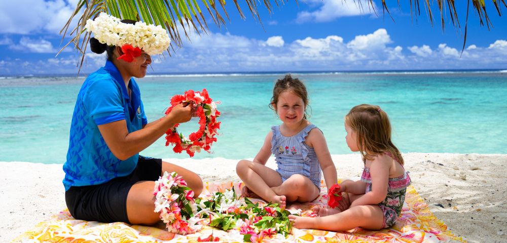 A woman in a blue shirt and floral headpiece making leis on a tropical beach with two young girls. They sit on a blanket under palm fronds, smiling.