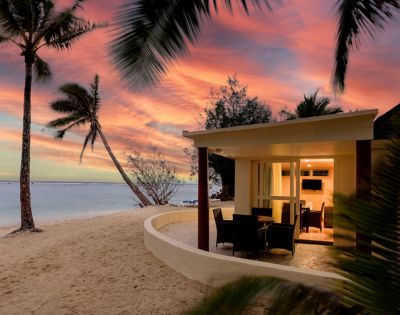 Beachfront bungalow at sunset with warm interior lights, surrounded by sand and palm trees. Vibrant orange and pink sky over a calm sea, evoking tranquility.