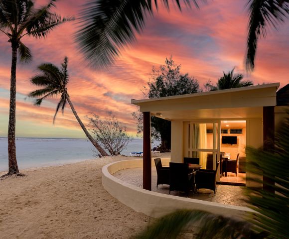 Beachfront bungalow at sunset with warm interior lights, surrounded by sand and palm trees. Vibrant orange and pink sky over a calm sea, evoking tranquility.