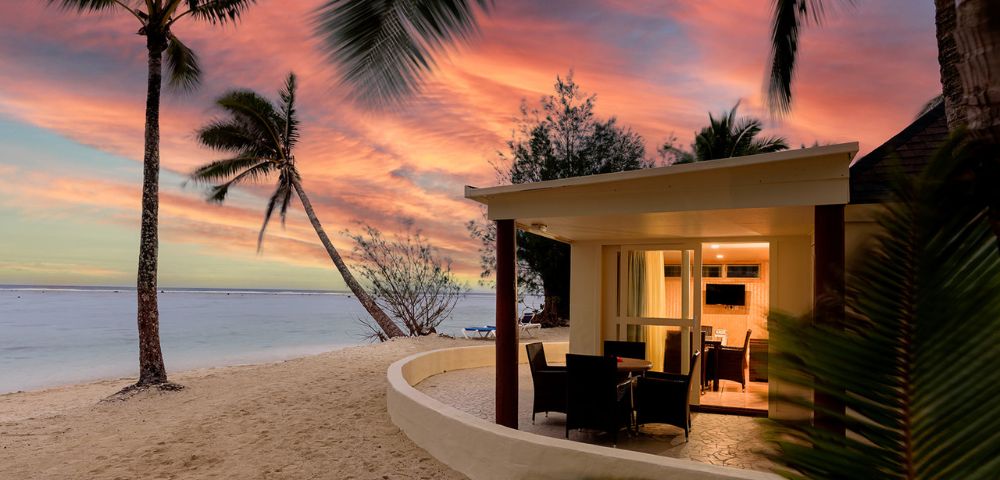 Beachfront bungalow at sunset with warm interior lights, surrounded by sand and palm trees. Vibrant orange and pink sky over a calm sea, evoking tranquility.