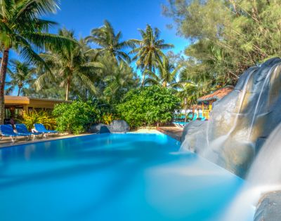 Tropical scene with a serene pool, gentle waterfall, and lush palm trees under a clear blue sky. Lounge chairs line the poolside, evoking relaxation.