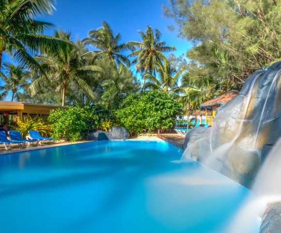 Tropical scene with a serene pool, gentle waterfall, and lush palm trees under a clear blue sky. Lounge chairs line the poolside, evoking relaxation.