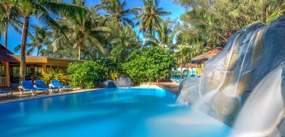 Tropical scene with a serene pool, gentle waterfall, and lush palm trees under a clear blue sky. Lounge chairs line the poolside, evoking relaxation.