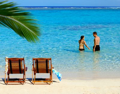 Two wooden lounge chairs on a sandy beach face a tranquil turquoise ocean. A couple stands in the shallow water under a clear blue sky.