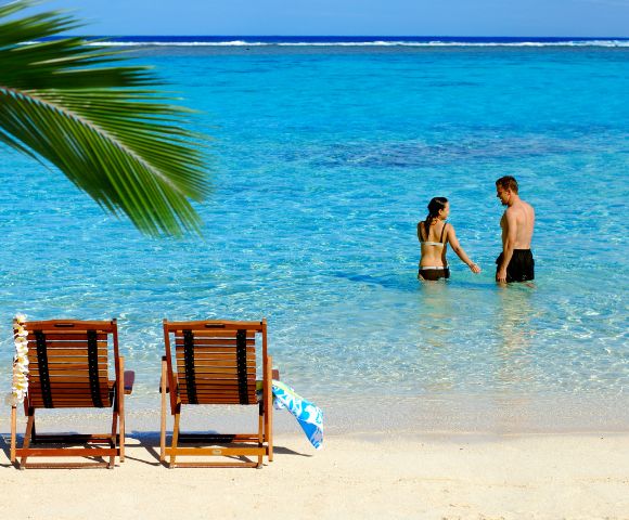 Two wooden lounge chairs on a sandy beach face a tranquil turquoise ocean. A couple stands in the shallow water under a clear blue sky.