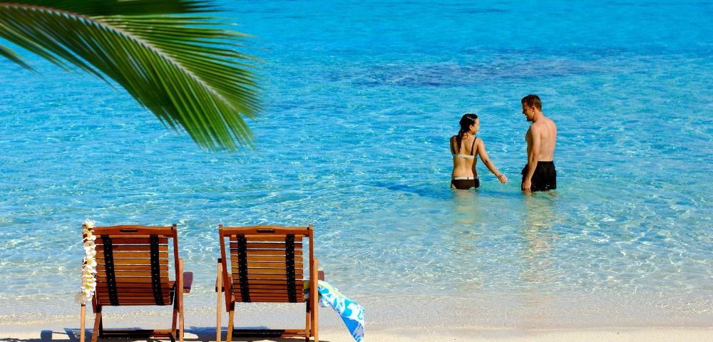 Two wooden lounge chairs on a sandy beach face a tranquil turquoise ocean. A couple stands in the shallow water under a clear blue sky.